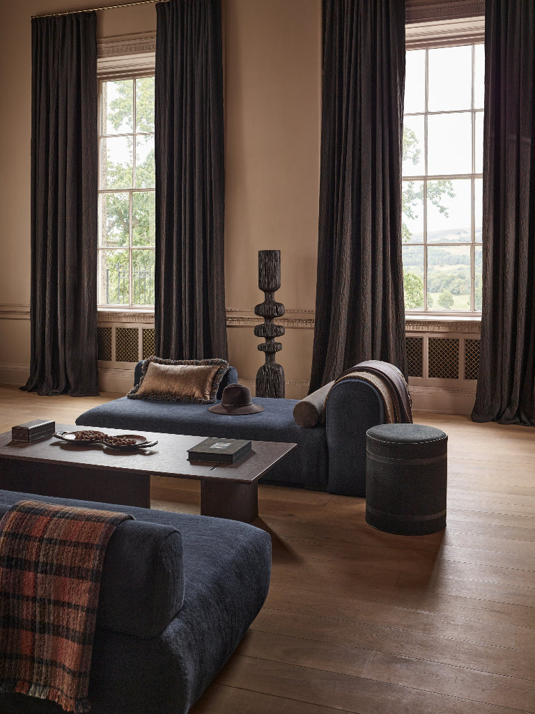 A styled interior room with dark charcoal gray curtains, a blue-gray chaise lounge with cushions, a matching coffee table, and a round ottoman on a wood floor.