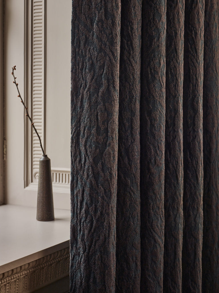 Dark textured window curtain hanging beside a window with a vase on the sill.