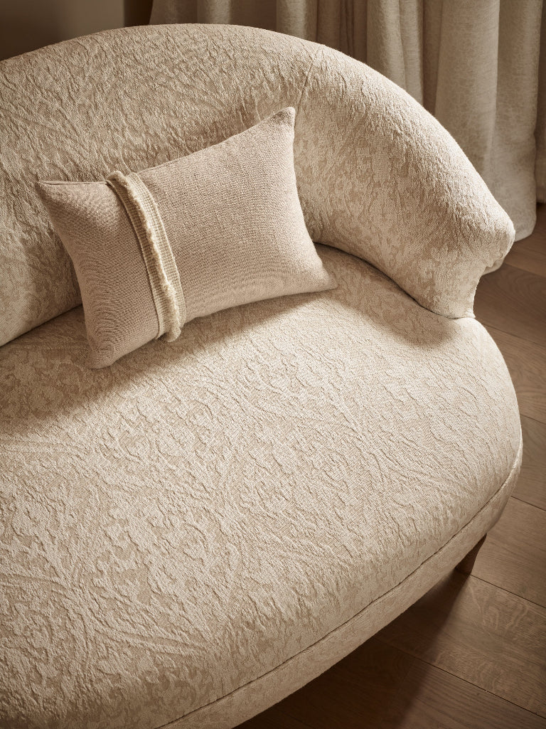 A close-up of a cream-colored upholstered armchair with a textured, embossed damask pattern, styled with a matching square throw pillow featuring a vertical ribbed stripe.