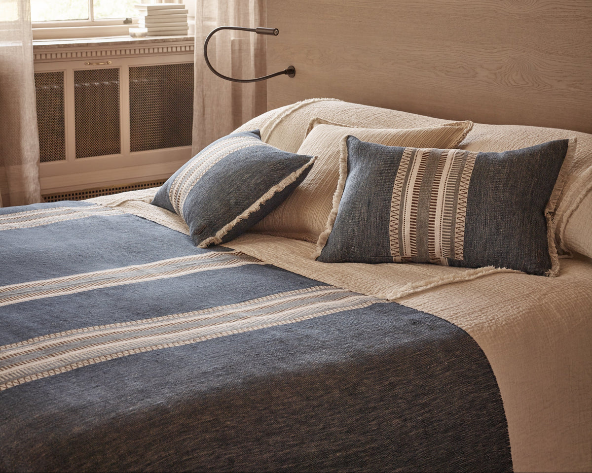 A styled bed in a warm bedroom setting featuring a textured slate blue throw blanket with cream stripes, matching blue and beige pillows, and a beige textured bedspread.