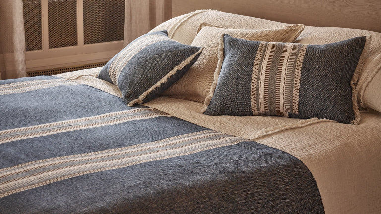 A styled bedroom scene featuring a bed dressed in beige linens with a slate blue striped throw blanket and matching pillows, set against a light wood headboard in a warm neutral room.