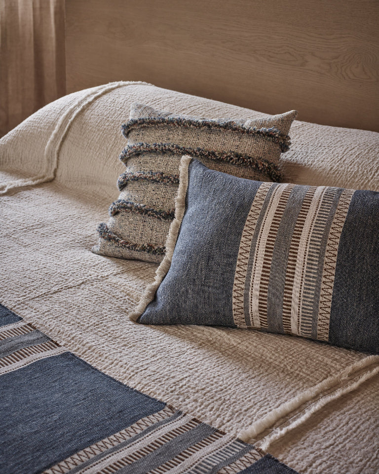 A styled bedroom scene showing textured throw pillows on a bed. The pillows include a large slate blue pillow with cream geometric stripes, a smaller striped beige and blue pillow, and a chunky cream bouclé pillow with fringed edges.