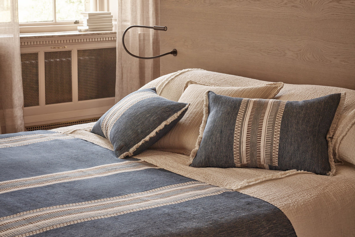 A styled bedroom scene featuring a bed dressed in beige linens with a slate blue striped throw blanket and matching pillows, set against a light wood headboard in a warm neutral room.
