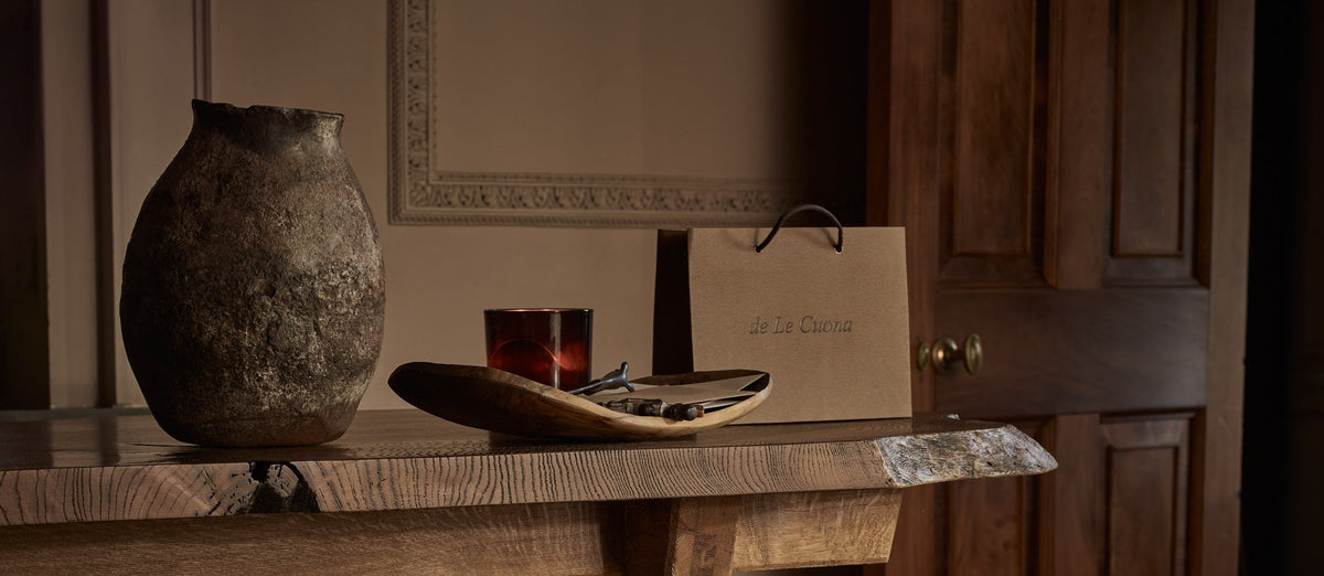 Two brown paper shopping bags with black handles and the printed brand name 'de La Couture' are displayed on a stone floor in a rustic, warm-toned room. A wooden table with a textured vase and other decorative items sits in the background.