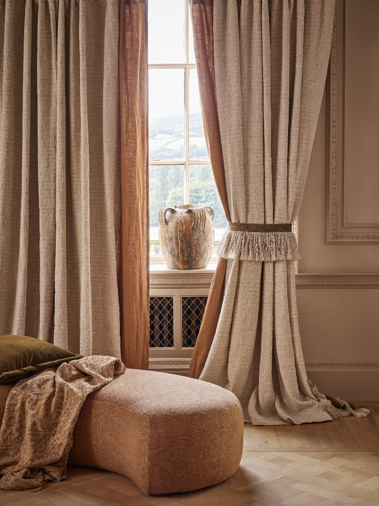 A styled interior scene showing floor-length curtains in warm beige and tan tones hanging around a window, with a matching upholstered ottoman in the foreground and a vase on the windowsill.