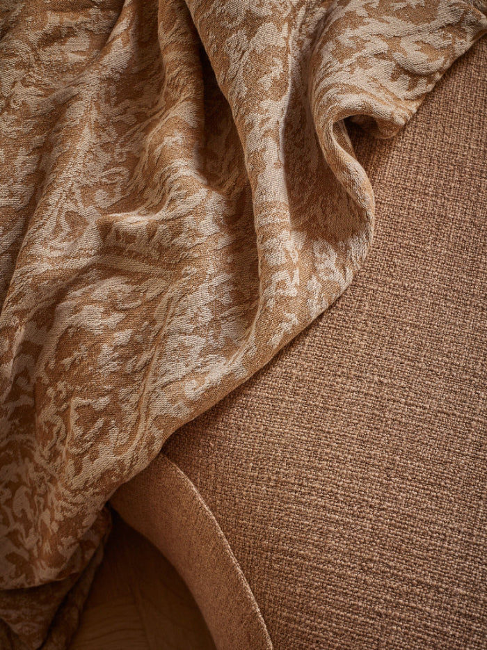 A close-up of draped fabric in warm beige and tan tones, showing a patterned textile layered over a textured brown surface.