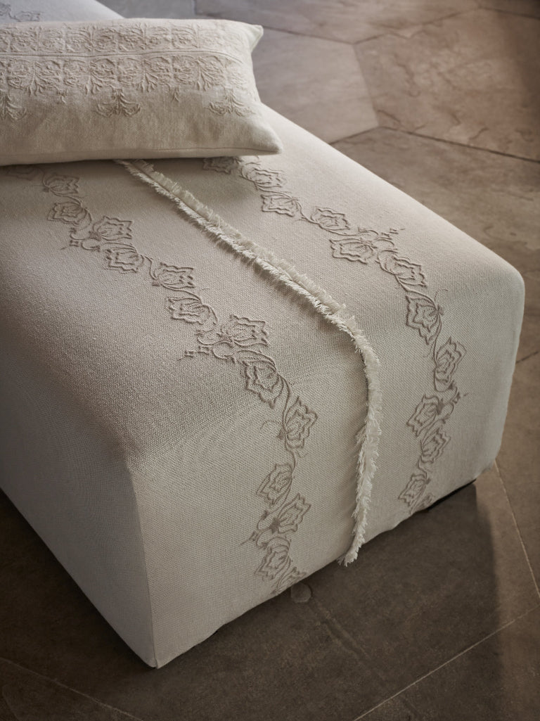 An off-white upholstered ottoman or bench with embroidered floral detailing and a fringed runner, shown in a styled interior setting with a matching pillow and stone tile flooring.