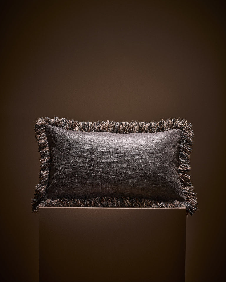 A rectangular dark brown textured throw pillow with fringed edges, displayed on a matching dark pedestal against a dark brown background.